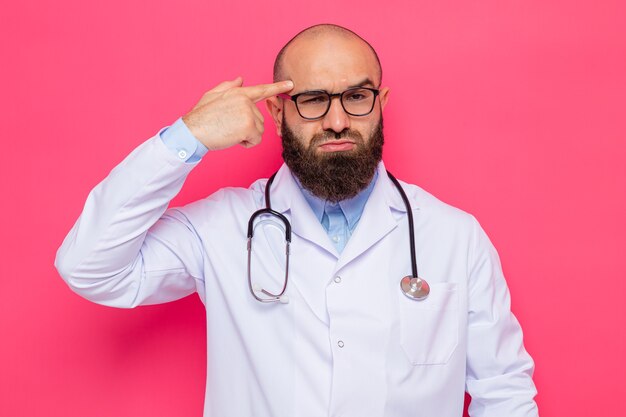 Bärtiger Arzt im weißen Kittel mit Stethoskop um den Hals mit Brille, der die Kamera mit stirnrunzelndem Gesicht betrachtet, das mit dem Zeigefinger auf seine Schläfe zeigt, die über rosafarbenem Hintergrund steht