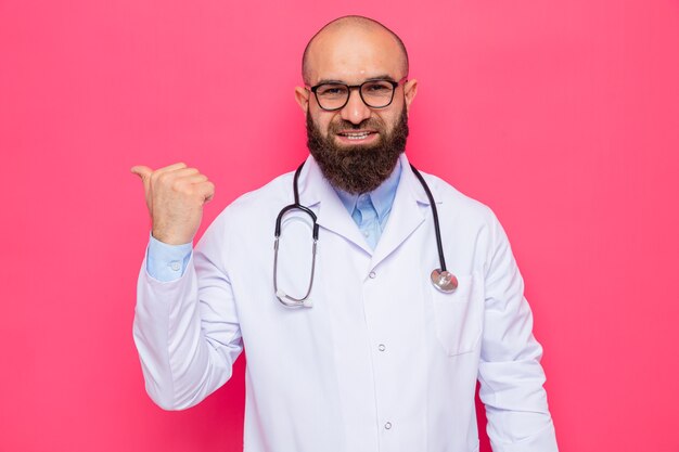 Bärtiger Arzt im weißen Kittel mit Stethoskop um den Hals mit Brille und Blick in die Kamera, die fröhlich lächelt und mit dem Daumen nach hinten auf rosafarbenem Hintergrund zeigt