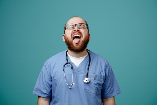 Kostenloses Foto bärtiger arzt in uniform mit stethoskop um den hals, der eine brille trägt, die die zunge herausstreckt, mit verärgertem gesichtsausdruck, der über blauem hintergrund steht