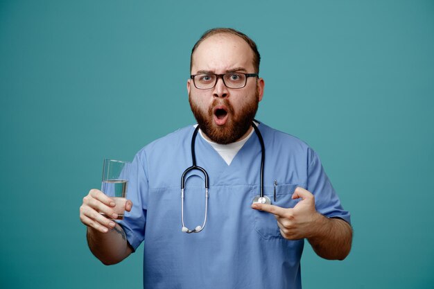 Bärtiger Arzt in Uniform mit Stethoskop um den Hals, der eine Brille trägt, die ein Glas Wasser hält und in die Kamera schaut, erstaunt und überrascht, wie er über blauem Hintergrund steht