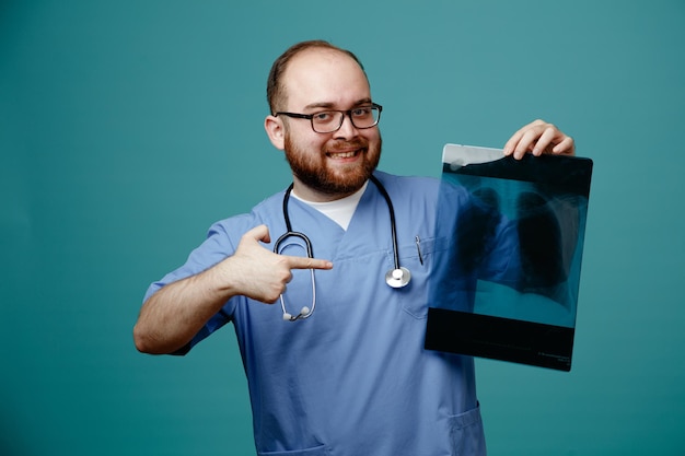 Bärtiger Arzt in Uniform mit Stethoskop um den Hals, der eine Brille trägt, die Lungenröntgen hält, glücklich und positiv mit dem Zeigefinger darauf zeigt und lächelnd über blauem Hintergrund steht