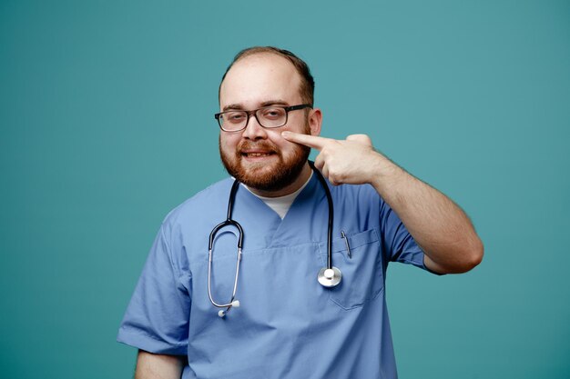Bärtiger Arzt in Uniform mit Stethoskop um den Hals, der eine Brille trägt und in die Kamera schaut, selbstbewusst lächelt und mit dem Zeigefinger auf seine Wange zeigt, die über blauem Hintergrund steht