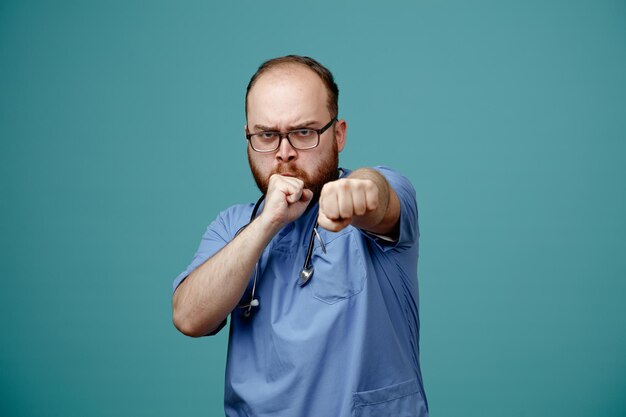 Bärtiger Arzt in Uniform mit Stethoskop um den Hals, der eine Brille trägt und mit ernsthaftem Gesicht, das die Fäuste ballt, in die Kamera blickt und sich wie ein Kämpfer aufstellt, der vor blauem Hintergrund steht