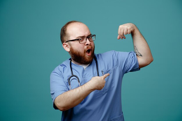 Bärtiger Arzt in Uniform mit Stethoskop um den Hals, der eine Brille trägt und selbstbewusst und stark die Faust hebt, die Bizeps zeigt, die über blauem Hintergrund stehen