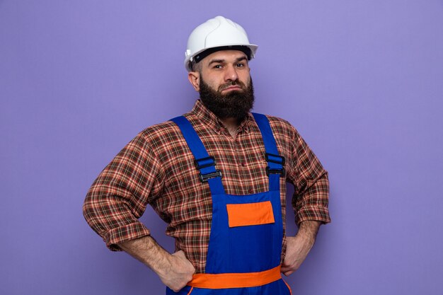 Bärtiger Baumeister in Bauuniform und Schutzhelm, der die Kamera mit selbstbewusstem Ausdruck mit den Händen an der Hüfte auf violettem Hintergrund anschaut