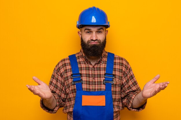 Bärtiger Baumeister in Bauuniform und Schutzhelm mit Blick auf die Kamera verwirrt, die Arme an den Seiten ausbreiten, die über orangefarbenem Hintergrund stehen