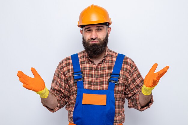 Bärtiger Baumeister in Bauuniform und Schutzhelm mit Gummihandschuhen, der verwirrt aussieht und die Arme an den Seiten ausbreitet und keine Antwort hat