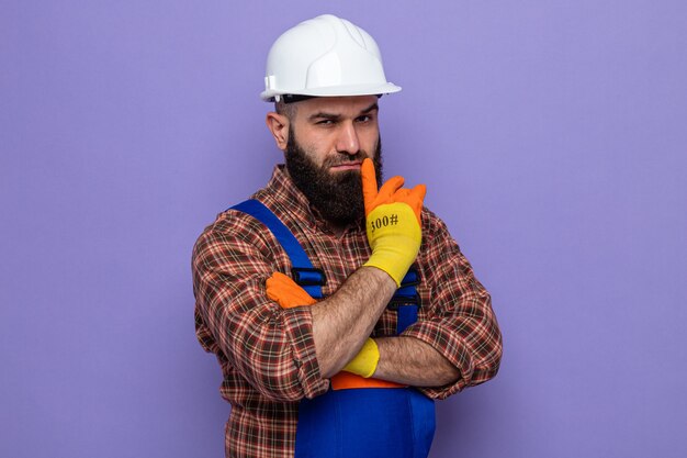 Bärtiger Baumeister in Bauuniform und Schutzhelm mit Gummihandschuhen, die mit ernsthaftem Gesichtsdenken aussehen