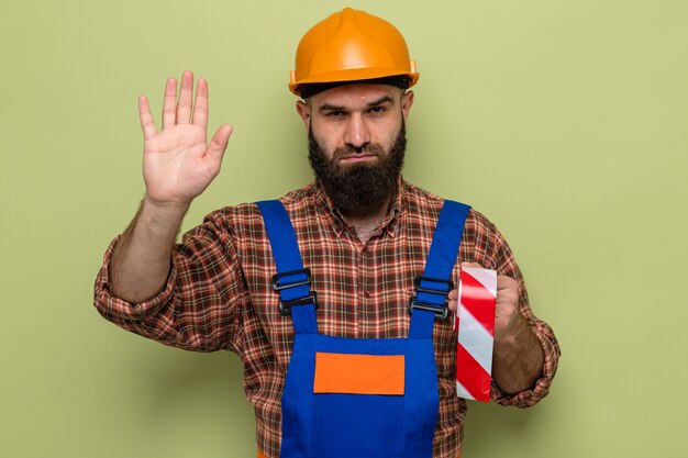 Bärtiger Baumeister in Bauuniform und Schutzhelm mit Klebeband, der mit ernstem Gesicht aussieht und mit der Hand eine Stopp-Geste macht