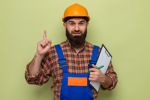 Bärtiger Baumeister in Bauuniform und Schutzhelm mit Klemmbrett mit Stift, der den Zeigefinger selbstbewusst lächelnd zeigt