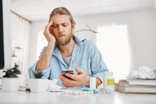 Bärtiger blonder männlicher Büroangestellter, der unglücklich auf den Bildschirm des Smartphones schaut, sich auf seinen Ellbogen stützt und während des harten Arbeitstages am Tisch vor dem Bildschirm sitzt. Manager leidet unter Kopfschmerzen.