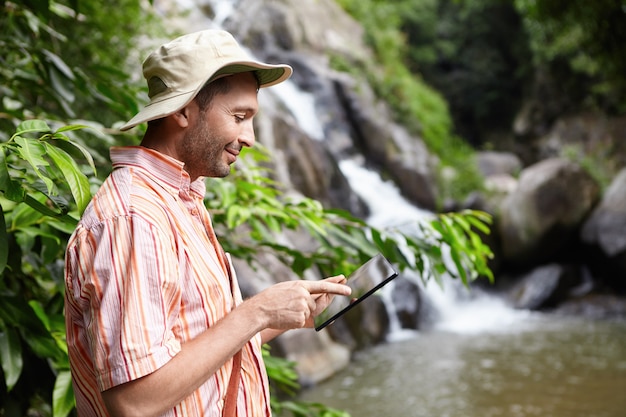 Bärtiger Botaniker, der Panamahut unter Verwendung der digitalen Tablette trägt, die auf leeren Bildschirm mit glücklichem Ausdruck zeigt und am Gebirgsfluss gegen Wasserfall steht, während Wasseranalyse durchführt