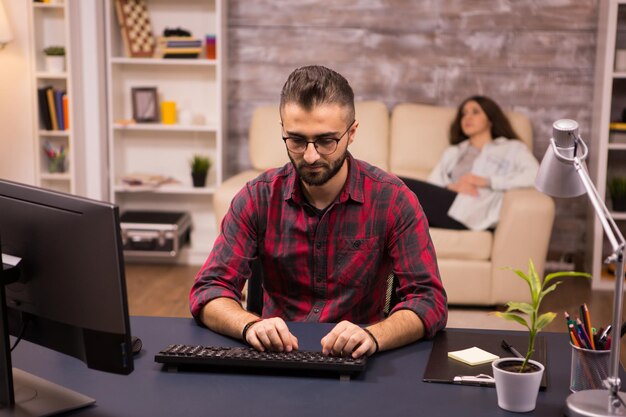 Bärtiger Freiberufler, der während der Arbeit von zu Hause aus am Computer tippt. Freundin entspannt auf dem Sofa im Hintergrund.