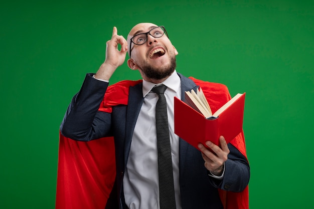 Bärtiger Geschäftsmann des Superhelden im roten Umhang, der offenes Buch hält, das oben zeigt Zeigefinger hat, der Idee lächelnd steht über grüner Wand