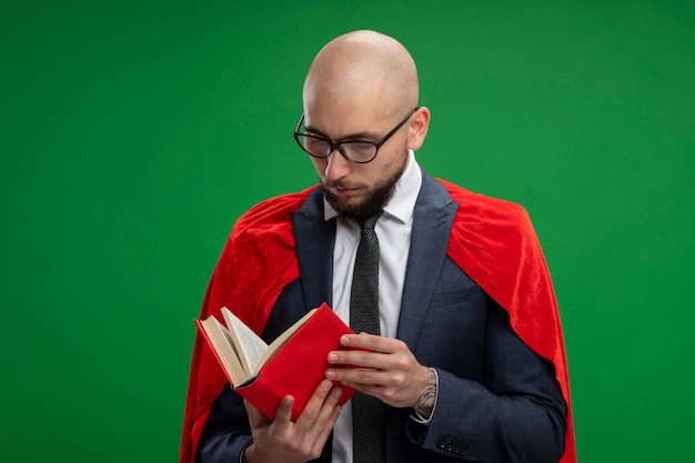 Bärtiger Geschäftsmann des Superhelden im roten Umhang, der offenes Buch liest, das über grüner Wand steht