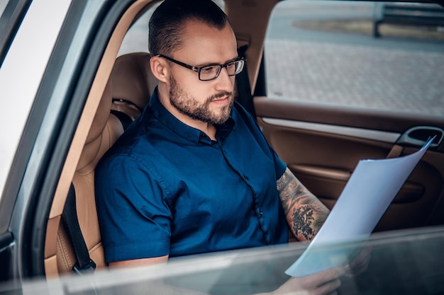 Bärtiger Geschäftsmann mit Brille und Tätowierung auf dem Arm sitzt auf dem Rücksitz eines Autos.