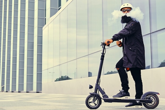 Bärtiger Hipster-Mann, der mit dem Elektroroller über modernes Gebäude dampft und fährt.