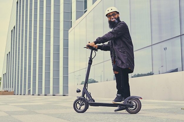 Bärtiger Hipster-Mann, der mit dem Elektroroller über modernes Gebäude dampft und fährt.