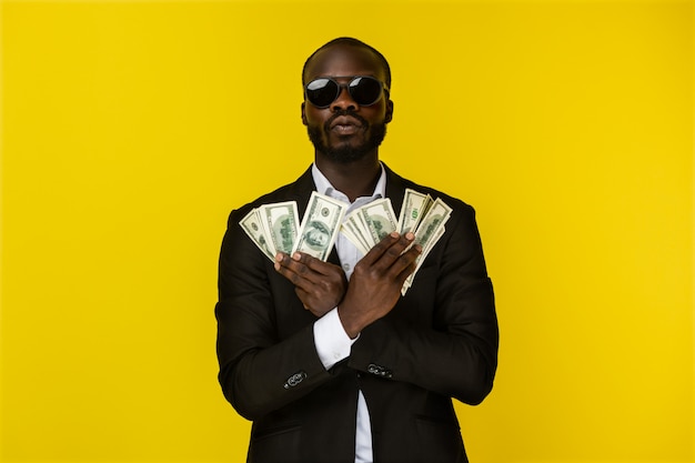 Bärtiger junger afroamerikanischer Luxuskerl hält viel Geld in beiden Händen in der Sonnenbrille und im schwarzen Anzug