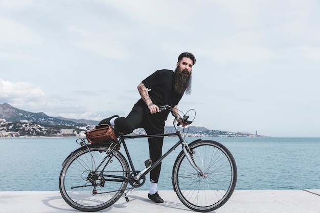 Bärtiger junger Mann, der auf Fahrrad nahe der Küste sitzt