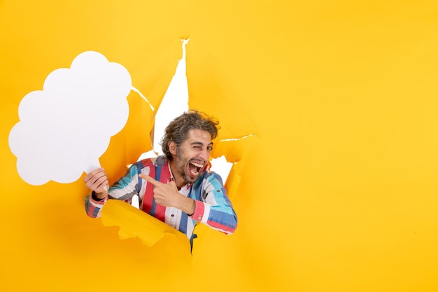 Bärtiger Kerl, der weißes wolkenförmiges Papier hält und etwas mit glücklichem Gesichtsausdruck in ein zerrissenes Loch und freien Hintergrund in gelbem Papier zeigt