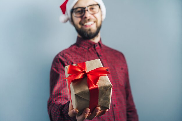 Bärtiger kerl in santa klausel hut und brille hält geschenk in bastelpapier eingewickelt und mit rotem sating-band in seinen händen verziert
