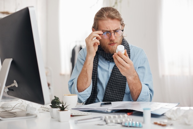 Bärtiger kranker männlicher Büroangestellter mit Brille auf liest Rezept der Medizin. Der junge Manager hat eine schlimme Erkältung und sitzt mit Pillen, Tabletten, Vitaminen und Drogen auf der Oberfläche am Tisch. Gesundheitsprobleme