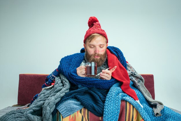 Bärtiger kranker Mann mit Kamin, der auf Sofa zu Hause oder im Studio mit Tasse Tee sitzt, der mit gestrickter warmer Kleidung bedeckt ist. Krankheit, Influenza-Konzept. Entspannung zu Hause. Gesundheitskonzepte.