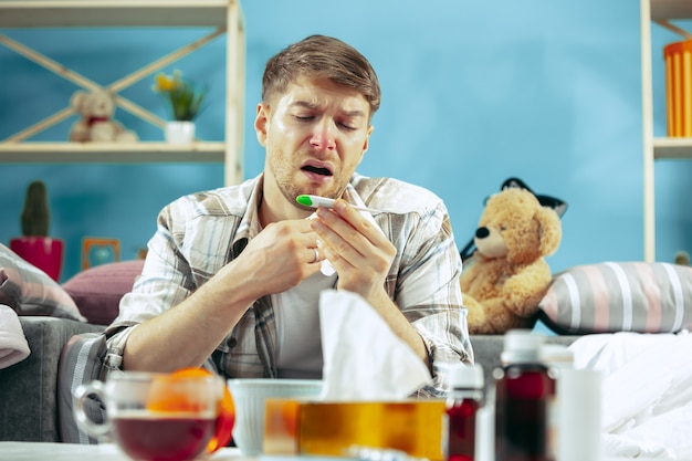 Kostenloses Foto bärtiger kranker mann mit kamin, der zu hause auf sofa sitzt und körpertemperatur misst. der winter, krankheit, influenza, schmerzkonzept. entspannung zu hause