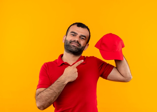 Bärtiger Lieferbote in der roten Uniform, die Kappe zeigt, zeigt mit dem Finger auf ihn lächelnd zuversichtlich stehend über orange Wand