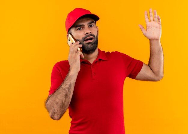 Bärtiger Lieferbote in der roten Uniform und in der Kappe, die auf Handy sprechen, der verwirrt und sehr ängstlich steht und über orange Wand steht