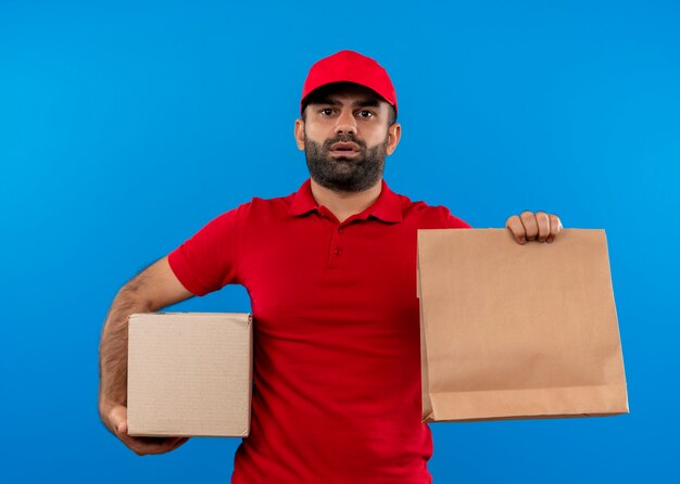 Bärtiger Lieferbote in der roten Uniform und in der Kappe, die Box und Papierpaket mit ernstem Gesicht hält, das über blauer Wand steht