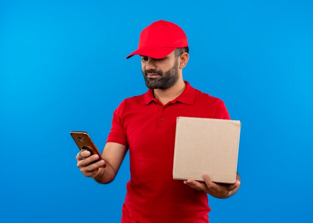 Bärtiger Lieferbote in der roten Uniform und in der Kappe, die Boxpaket hält, das Bildschirm seines Handys mit ernstem Gesicht betrachtet, das über blauer Wand steht