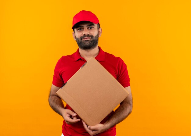 Bärtiger Lieferbote in der roten Uniform und in der Kappe, die Boxpaket mit Lächeln auf Gesicht über orange Wand hält