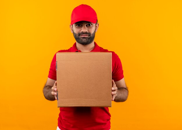 Bärtiger Lieferbote in der roten Uniform und in der Kappe, die Boxpaket mit sicherem ernstem Ausdruck hält, der über orange Wand steht
