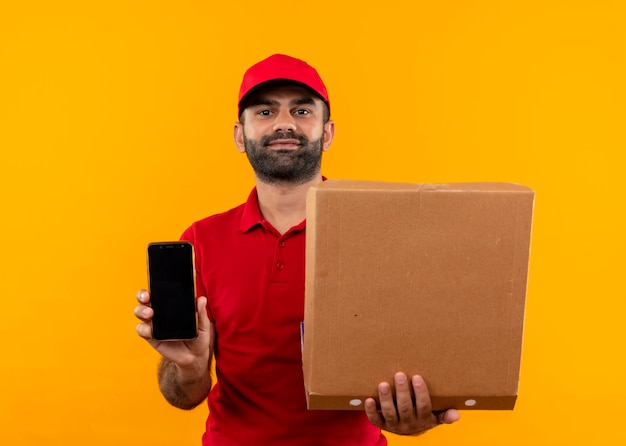 Bärtiger Lieferbote in der roten Uniform und in der Kappe, die die offene Pizzaschachtel zeigt, die Smartphone mit sicherem Ausdruck zeigt, der über orange Wand steht