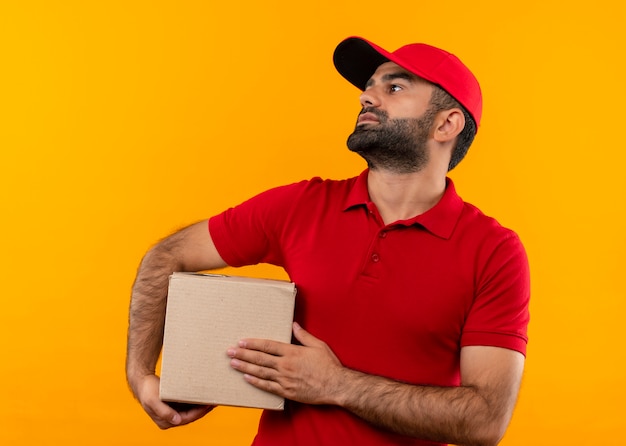 Bärtiger Lieferbote in der roten Uniform und in der Kappe, die Kastenpaket beiseite schaut mit ernstem Gesicht, das über orange Wand steht