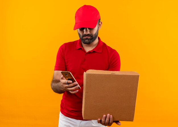 Bärtiger Lieferbote in der roten Uniform und in der Kappe, die offene Pizzaschachtel-SMS auf Handynachricht hält, die über orange Wand steht