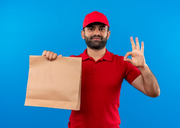 Bärtiger Lieferbote in der roten Uniform und in der Kappe, die Papierpaket lächelnd zeigt okes Zeichen, das über blauer Wand steht