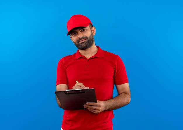 Bärtiger Lieferbote in der roten Uniform und in der Kappe, die Zwischenablage schreiben, die sicher steht, über blauer Wand zu stehen