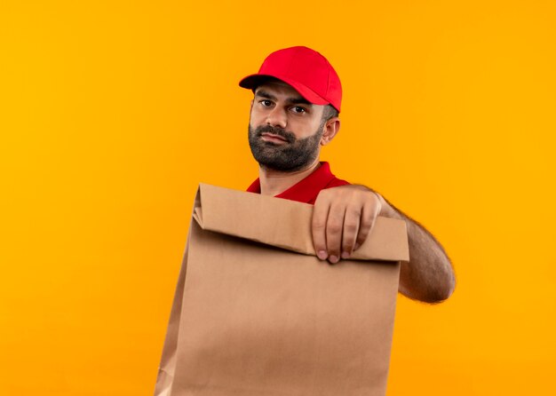 Bärtiger Lieferbote in roter Uniform und Kappe, die Papierpaket mit ernstem Gesicht hält, das über orange Wand steht