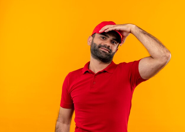 Bärtiger Lieferbote in roter Uniform und Mütze mit Hand auf seiner Kamera lächelnd über orange Wand stehend
