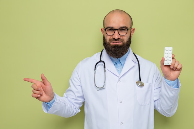 Bärtiger Mann Arzt im weißen Kittel mit Stethoskop um den Hals tragen Brille hält Blase mit Pillen glücklich und positiv mit Zeigefinger zur Seite lächelnd