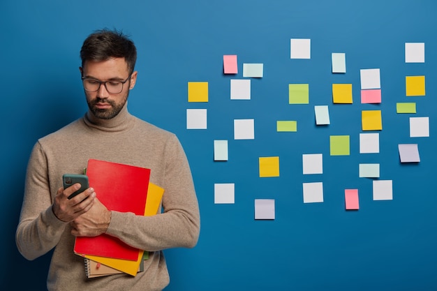 Kostenloses Foto bärtiger mann benutzt handy für online-konversation, hält lehrbuch, trägt eine brille, trägt eine brille und einen braunen pullover, sucht nach informationen, bunte notizen hinter der wand