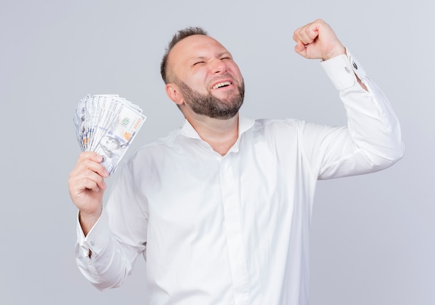Bärtiger Mann, der weißes Hemd hält, das Bargeld, das die Faust ballt, glücklich und aufgeregt freut, über weißer Wand stehend