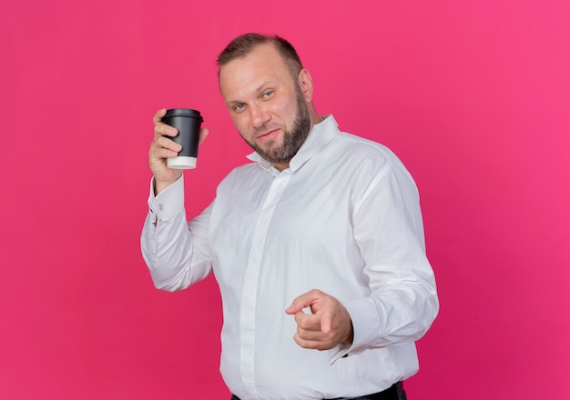 Bärtiger Mann, der weißes Hemd hält, das Kaffeetasse pointign mit lächelndem Zeigefinger hält, der über rosa Wand steht