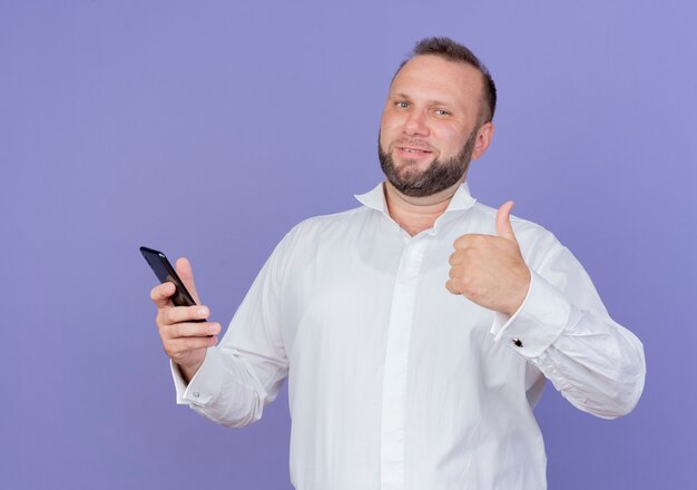 Bärtiger Mann, der weißes Hemd hält, das lächelndes Smartphone hält und Daumen hoch zeigt, die über blaue Wand stehen