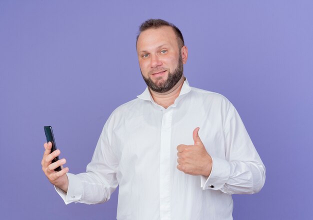 Bärtiger Mann, der weißes Hemd hält, das lächelndes Smartphone hält und Daumen hoch zeigt, die über blaue Wand stehen