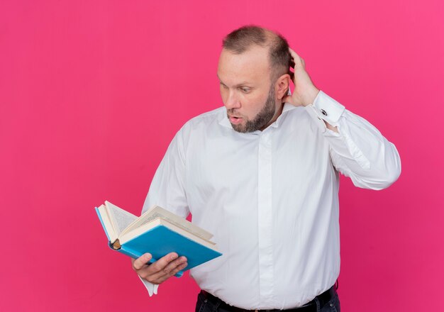 Bärtiger Mann, der weißes Hemd hält, das offenes Buch hält und es verwirrt betrachtet, über rosa Wand stehend