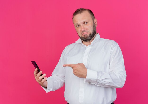 Bärtiger Mann, der weißes Hemd hält, das Smartphone hält, das mit Finger auf es schaut und sicher steht, über rosa Wand zu stehen